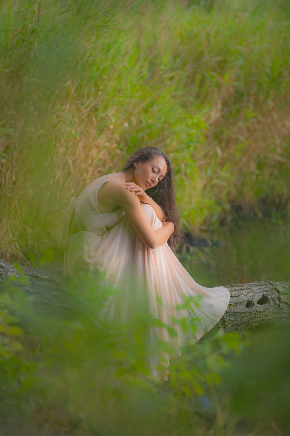 Ethereal figure in light dress sits dreamily on fallen log beside pond in soft natural lighting.