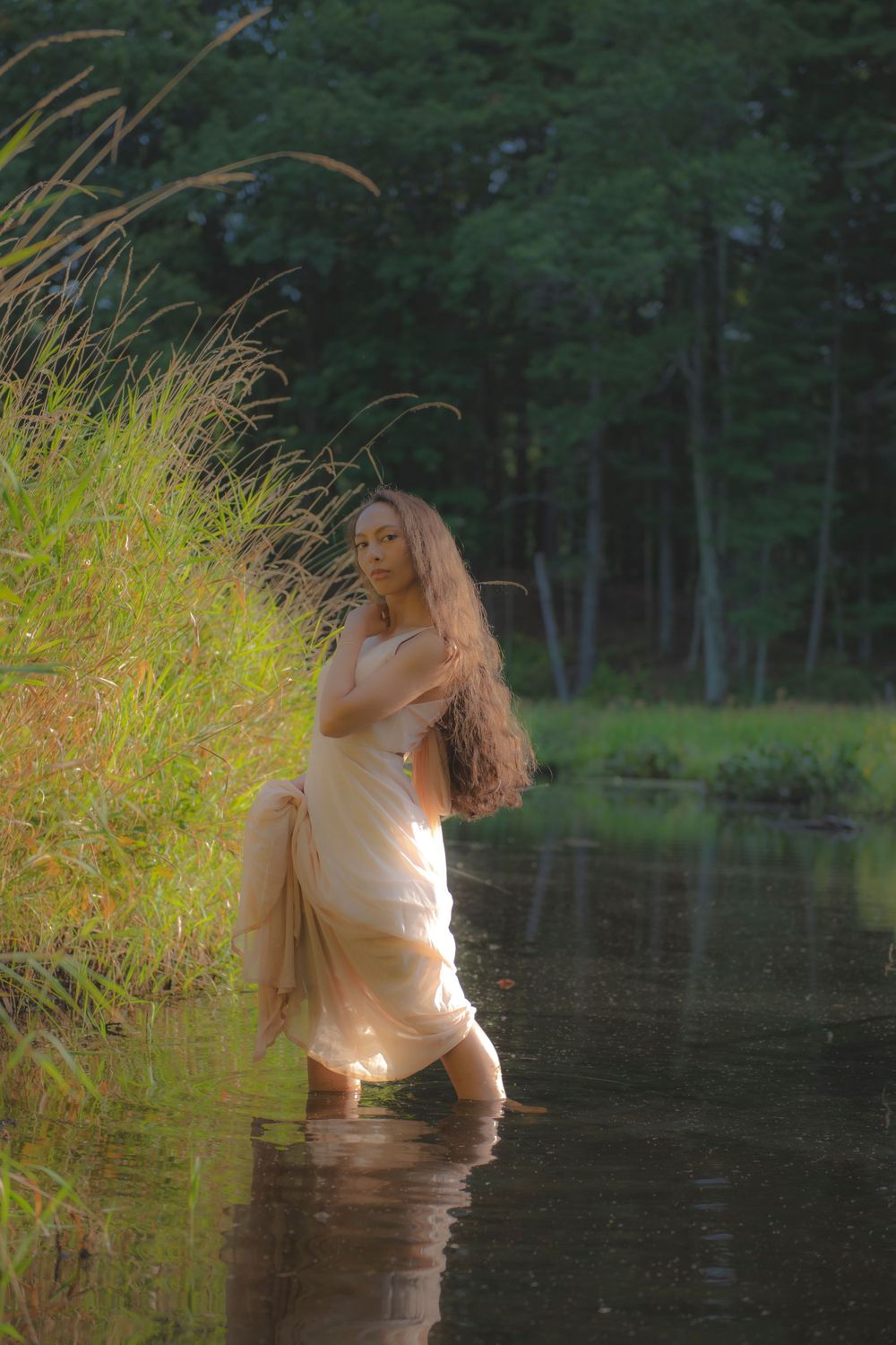 Flowing dress catches golden light as figure stands gracefully in shallow forest pond.