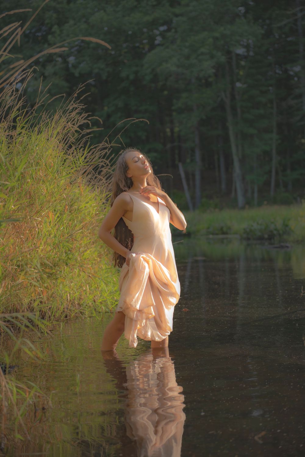 Artistic portrait of figure in cream dress standing in reflective pond waters at sunset.