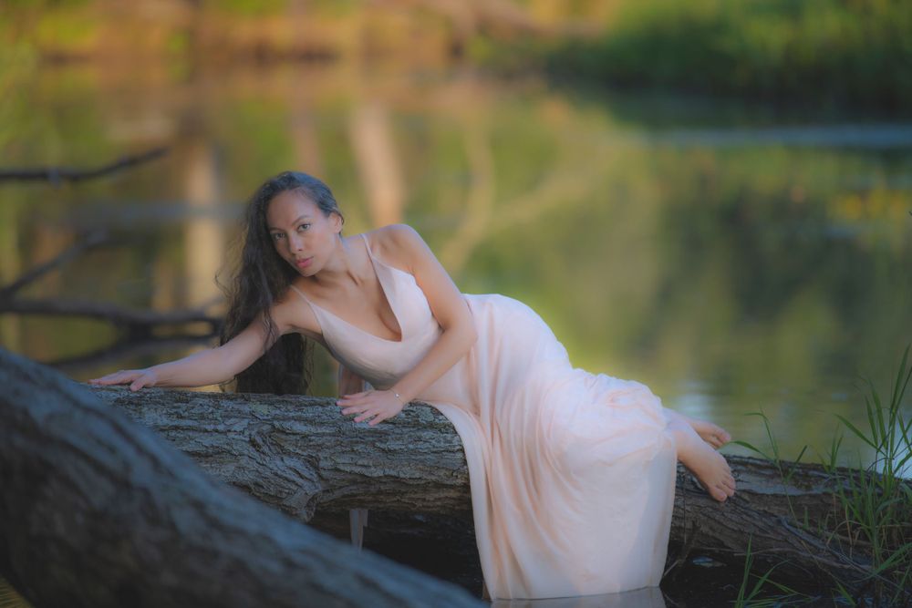 Ethereal portrait in a soft peach dress reclining on a fallen log by a tranquil pond at sunset.