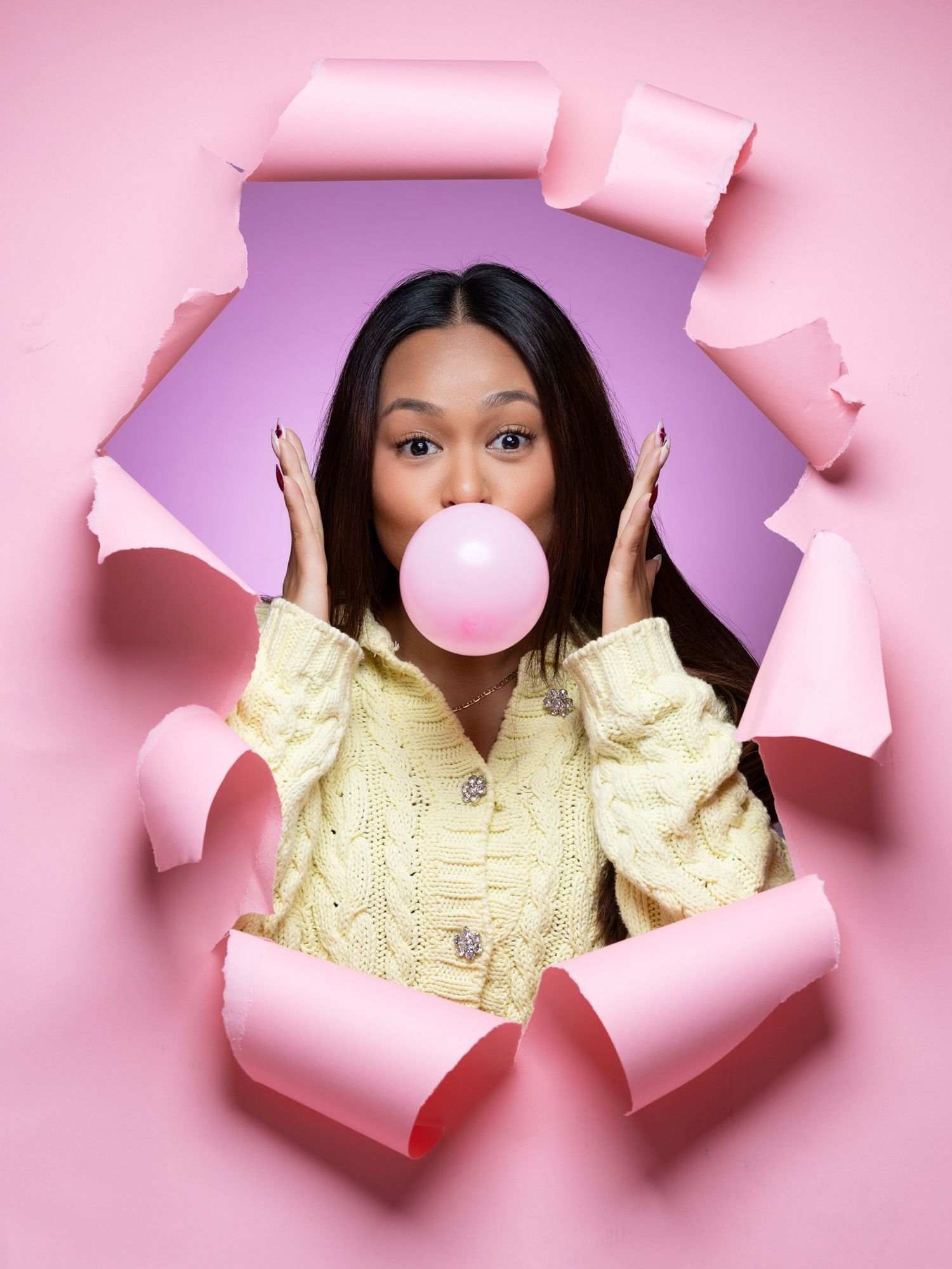 Creative portrait through torn pink paper showing a person blowing a pink bubble gum bubble wearing a yellow jacket.