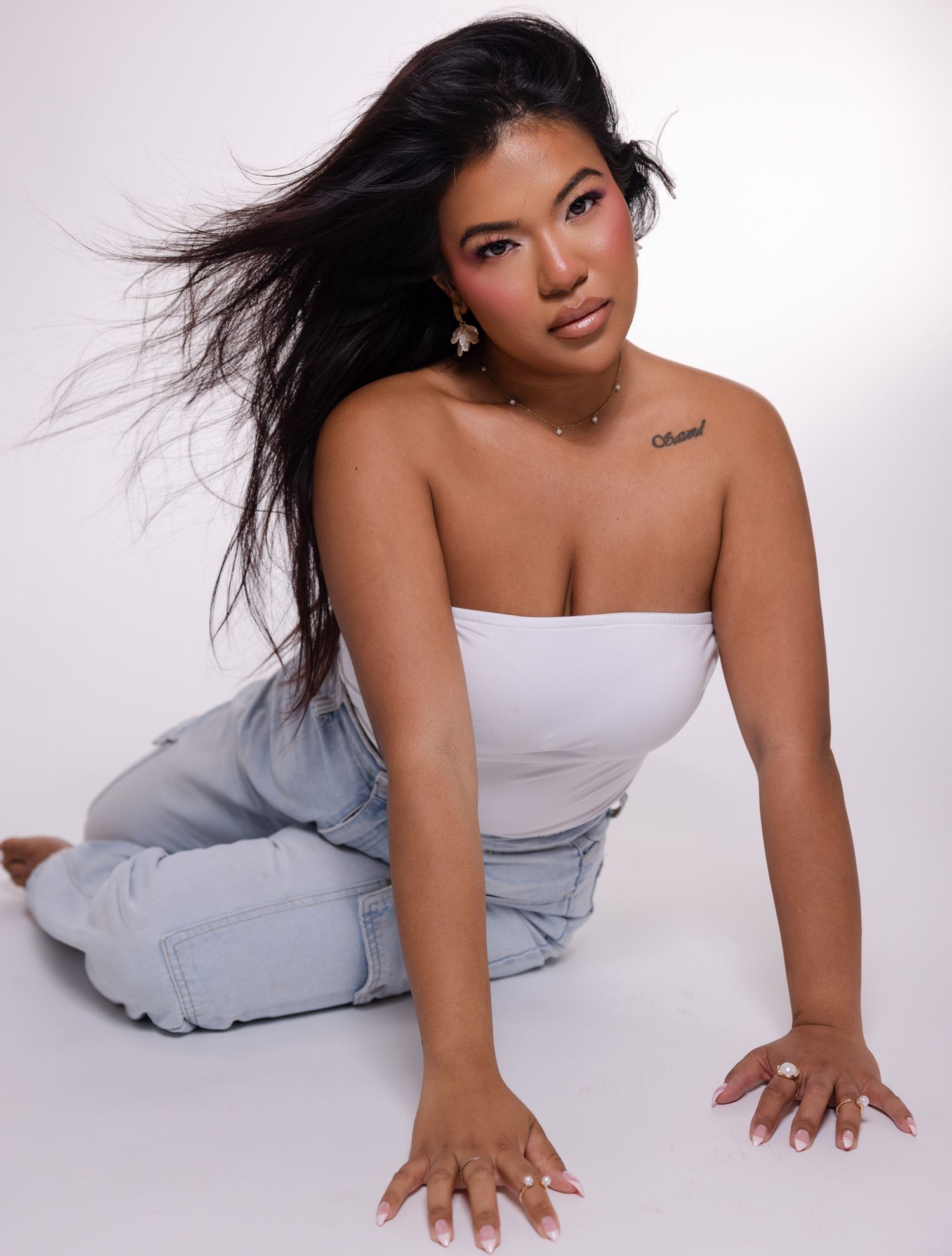 Model in white tube top and light jeans poses stylishly on studio floor with windswept black hair.