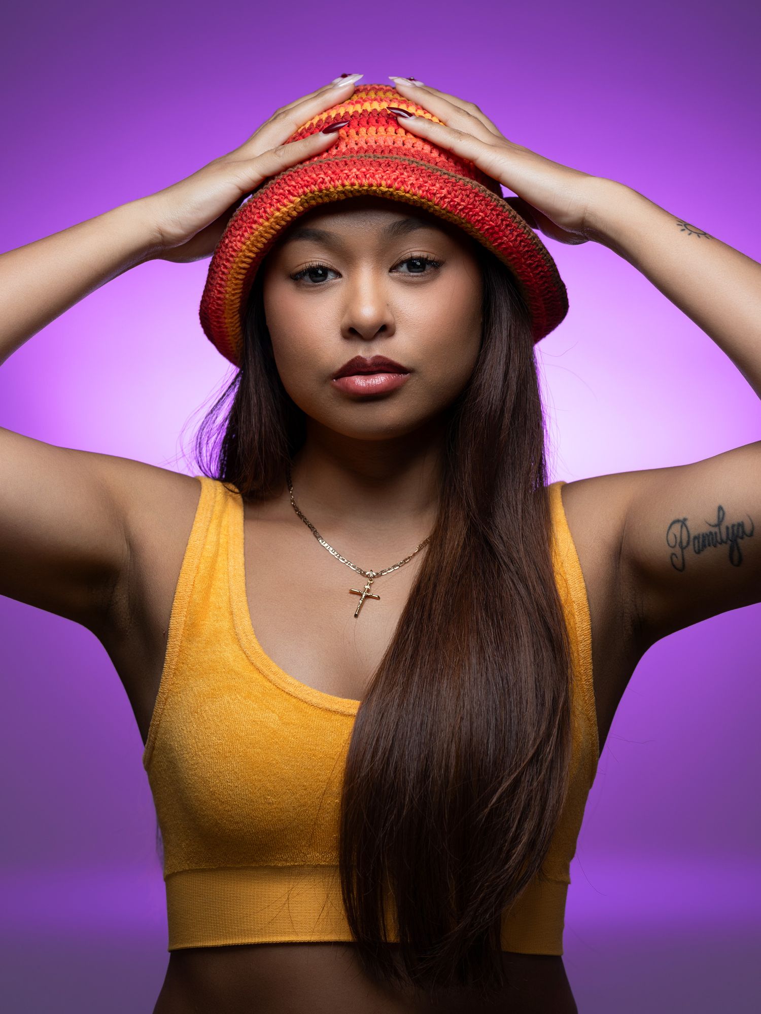 Fashion model in yellow tank top and red knit bucket hat poses against purple backdrop with long dark hair and gold necklace.