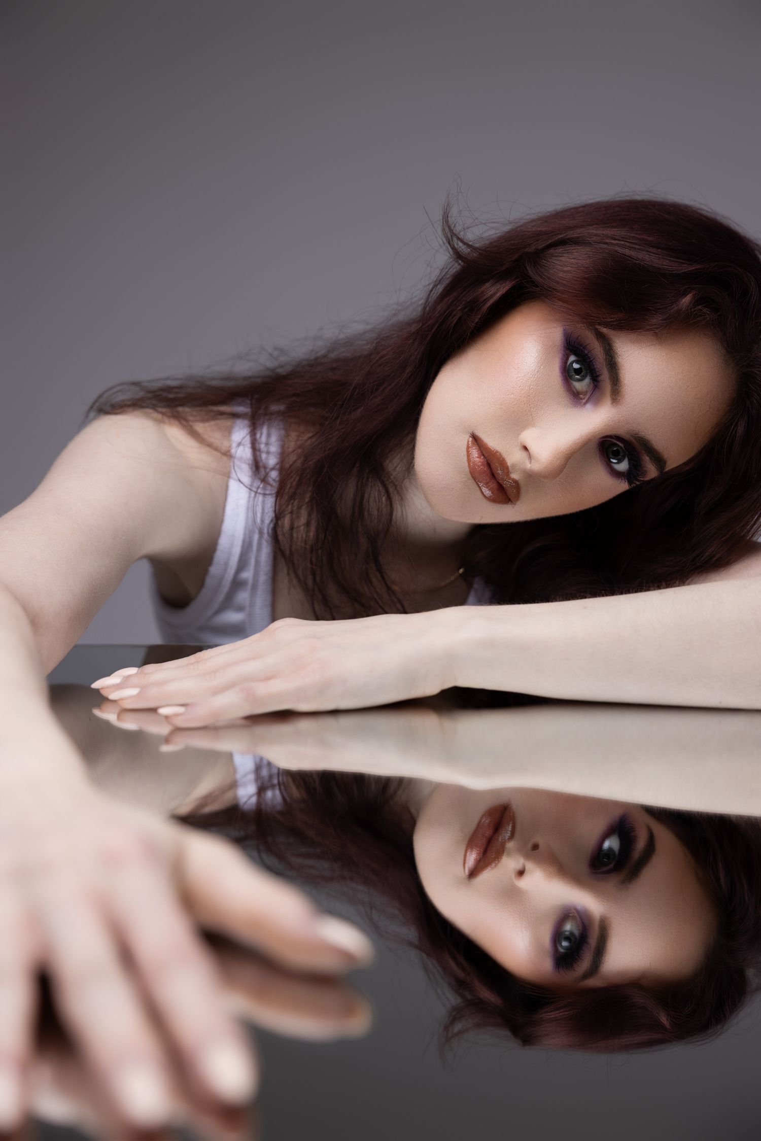 Artistic portrait with mirror reflection showing dark hair and dramatic makeup against grey background.