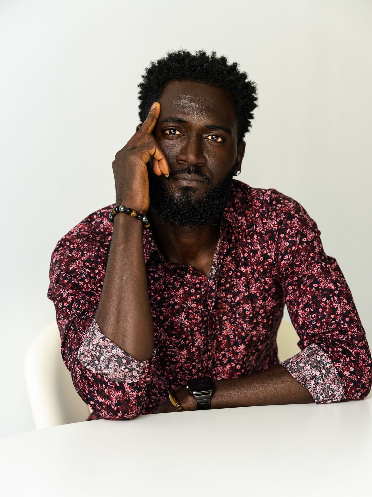 Male model in pink and burgundy floral patterned button-up shirt posing thoughtfully against white background.