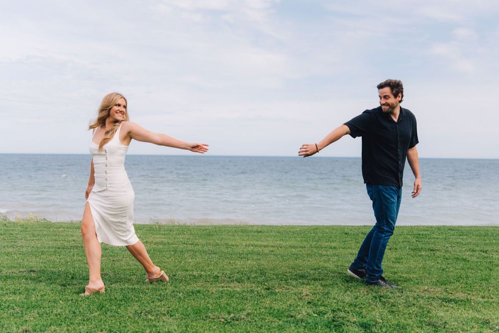 Playful couple reaching towards each other on grass by the ocean.