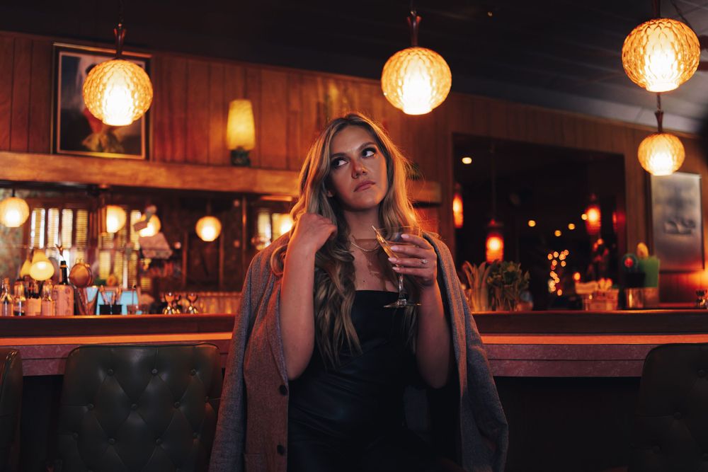 Woman in dark attire poses at a moody bar with warm lighting and vintage ambiance.