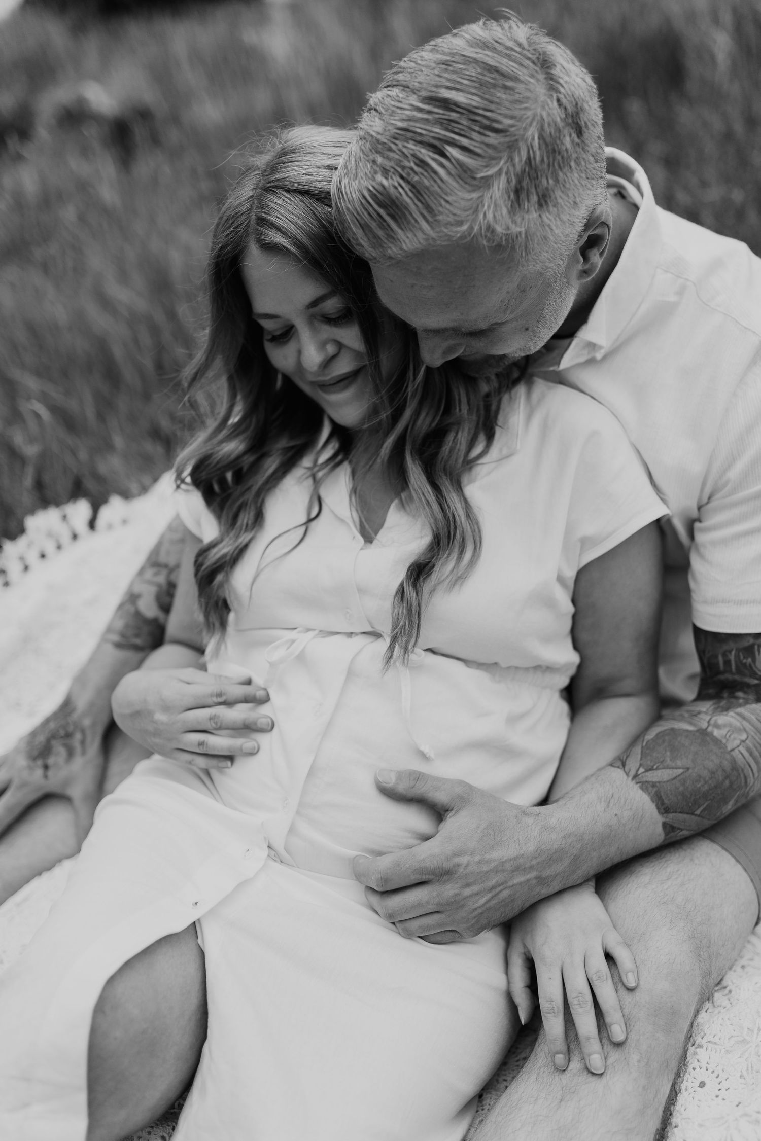 pregnant lady sitting in between her husband's legs, while he hugs her belly