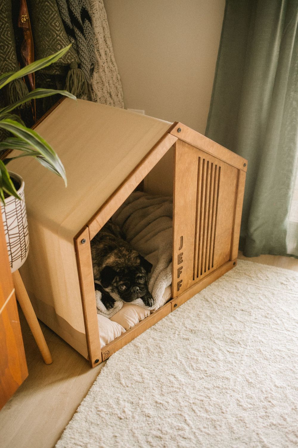 A black pug dog sleeping inside a modern wooden dog house with soft cushions in a stylish home interior.