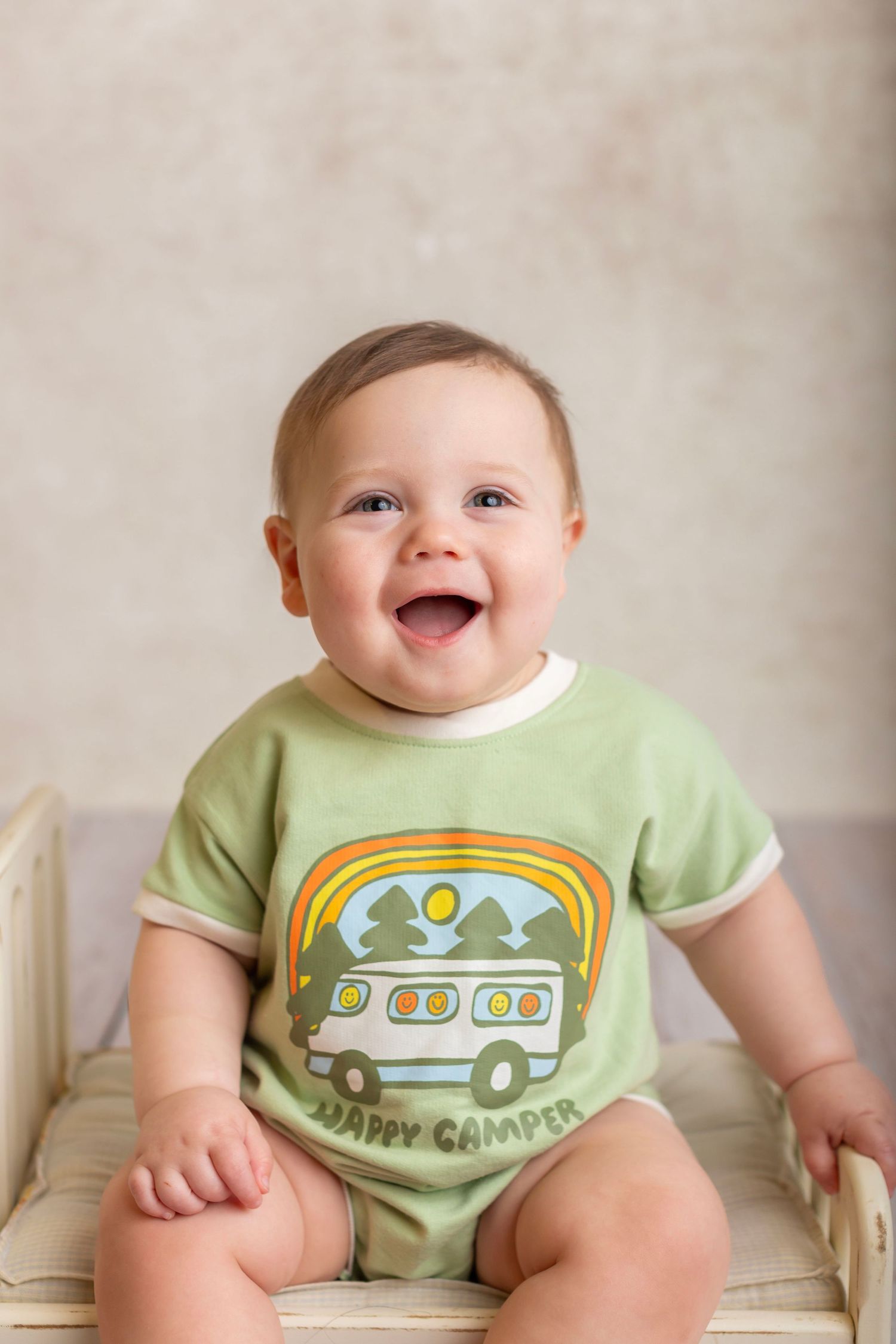 Laughing baby wearing green onesie with camping design sits in chair.