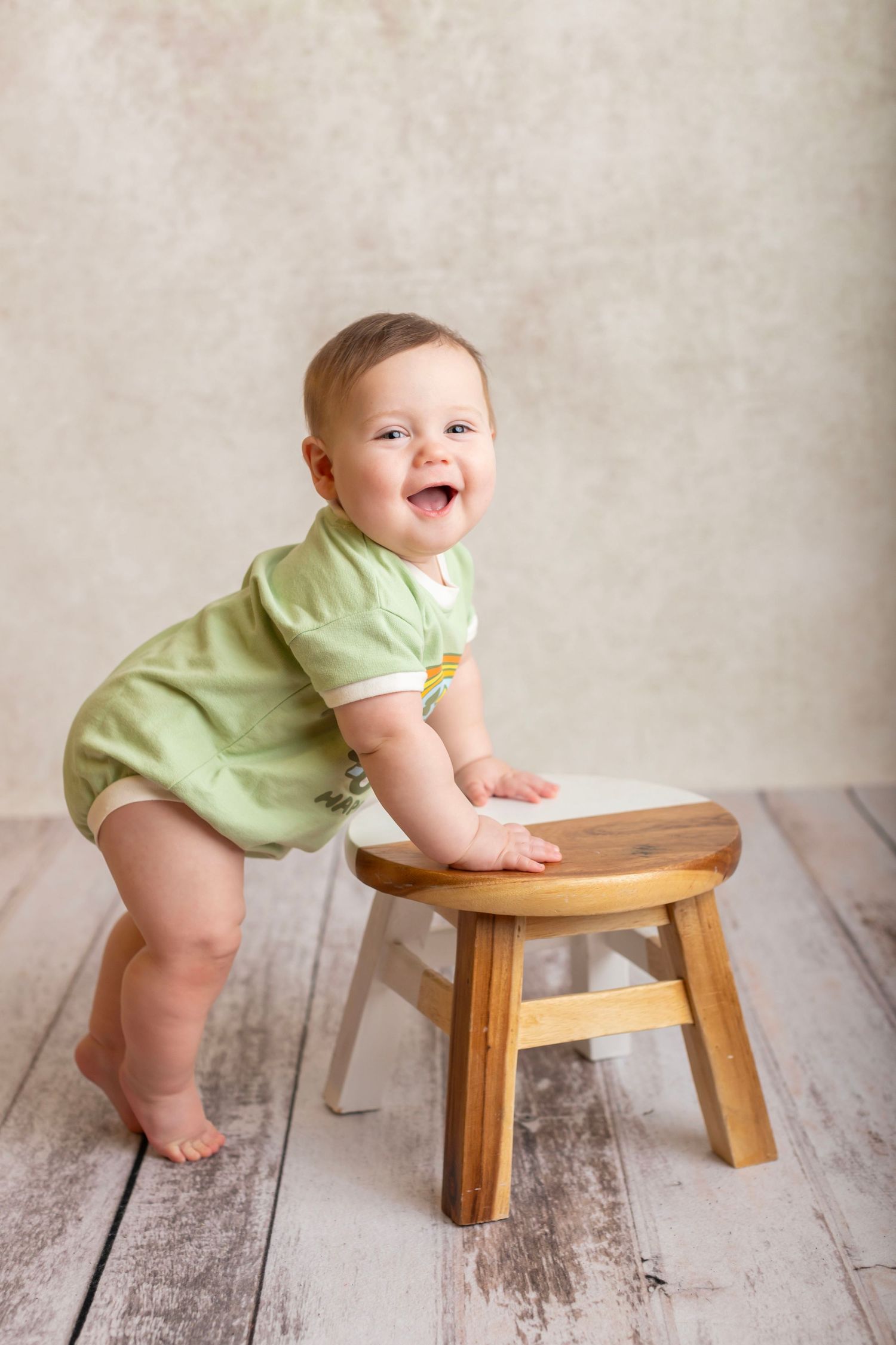 Happy baby in green onesie standing at wooden stool against neutral background.