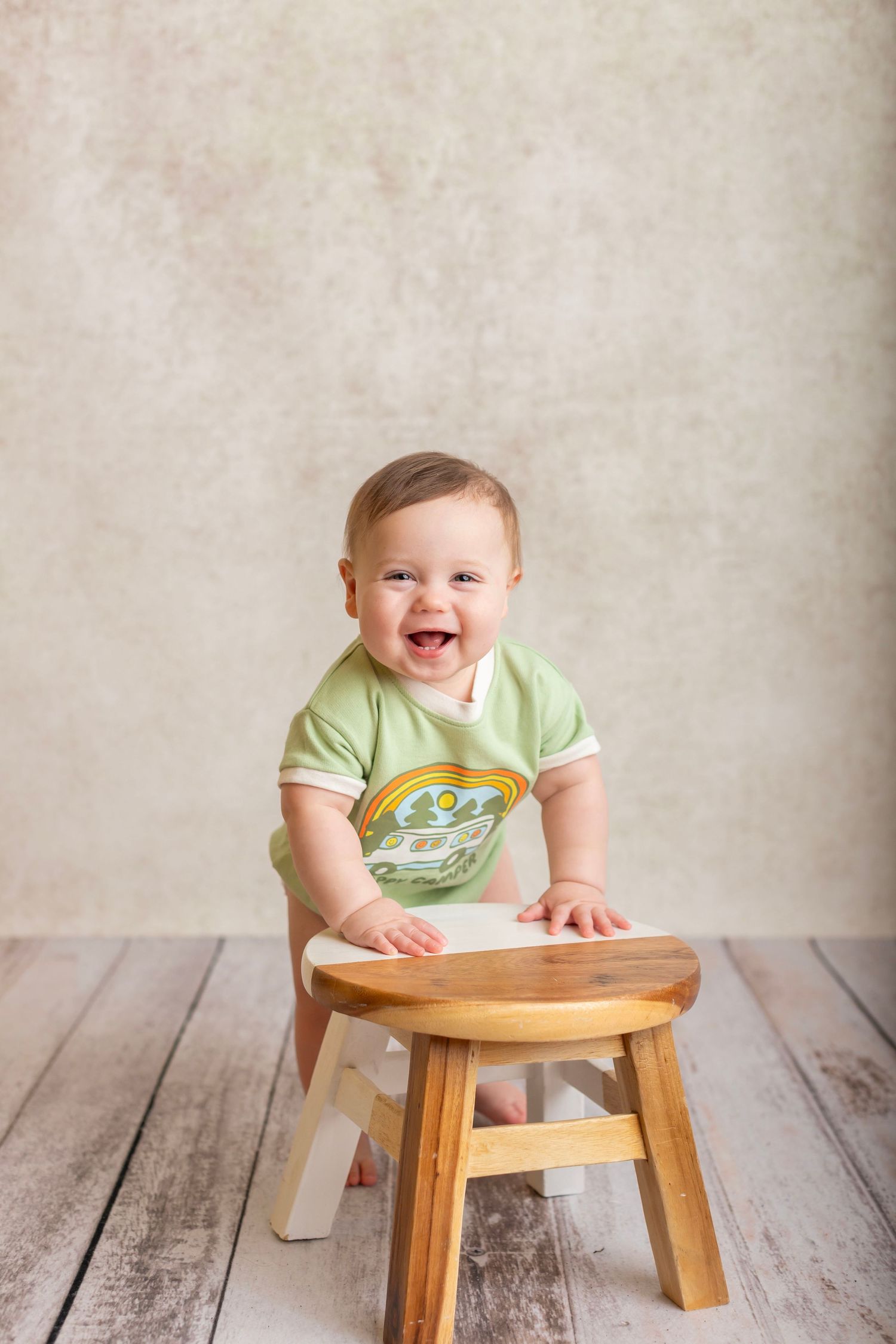 Baby wearing green shirt smiles joyfully while leaning on a wooden stool against light textured backdrop.