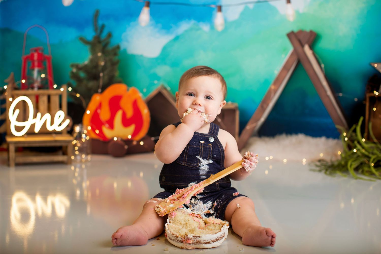 Baby with cake wearing navy outfit against teal backdrop with string lights and festive decor.