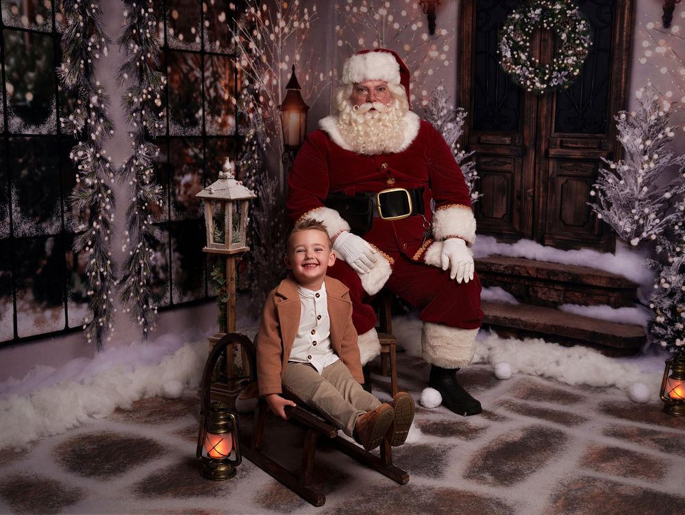 A festive holiday scene with Santa Claus and a small child in a brown coat sitting by a lantern in a snowy setting.