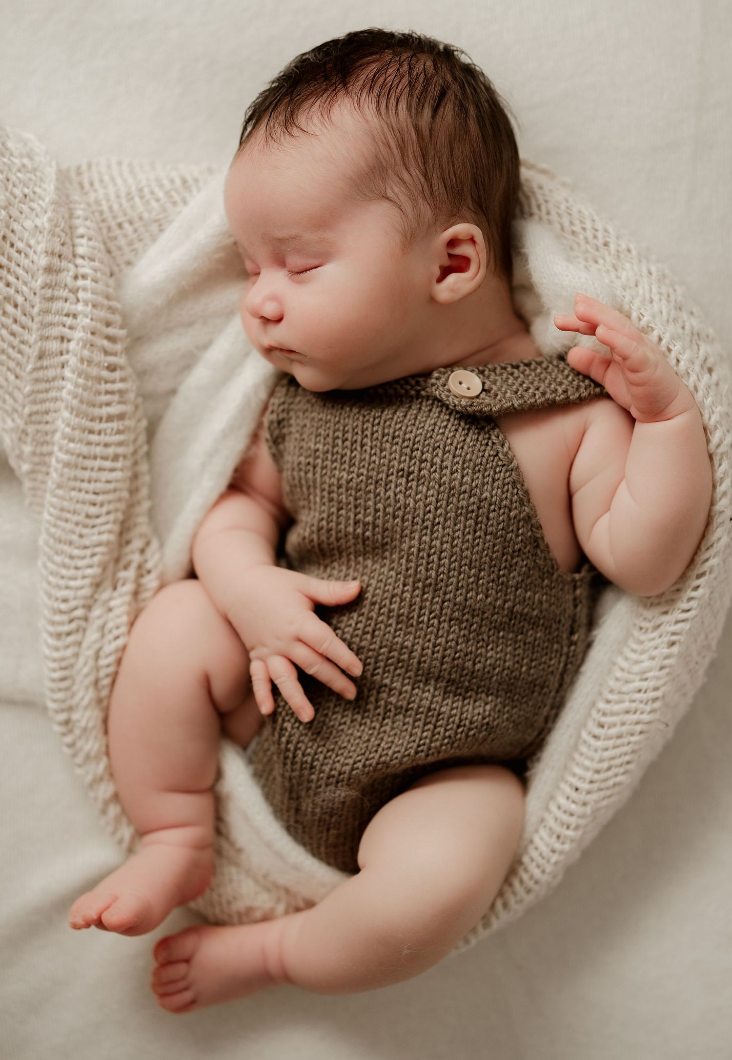 Sleeping newborn baby in olive green knit romper nestled in white knit wrap.
