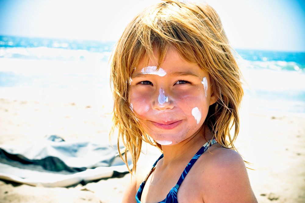 Child with sunscreen on nose smiles brightly on sunny beach day.
