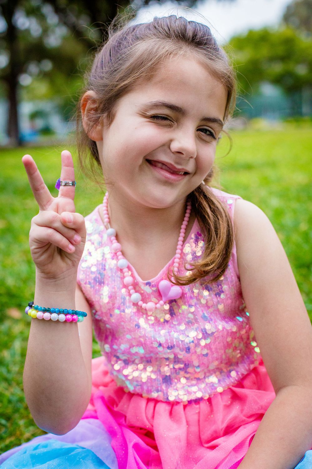 Child wearing sparkly pink dress and colorful bracelets showing peace sign while sitting outdoors in grassy area.