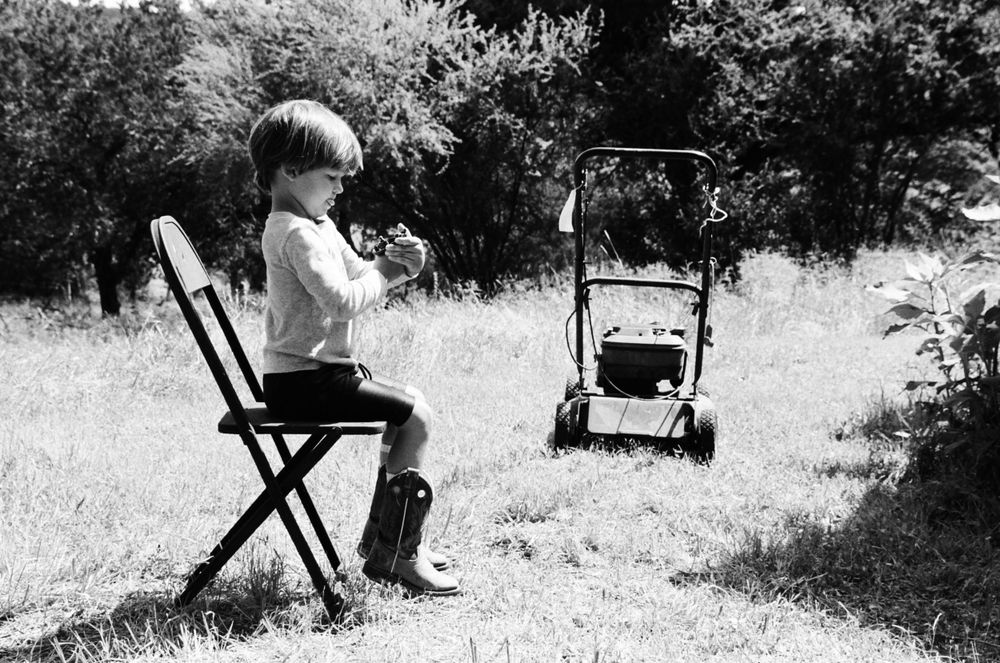 Black and white photo of child playing flute outdoors with lawn mower in background.
