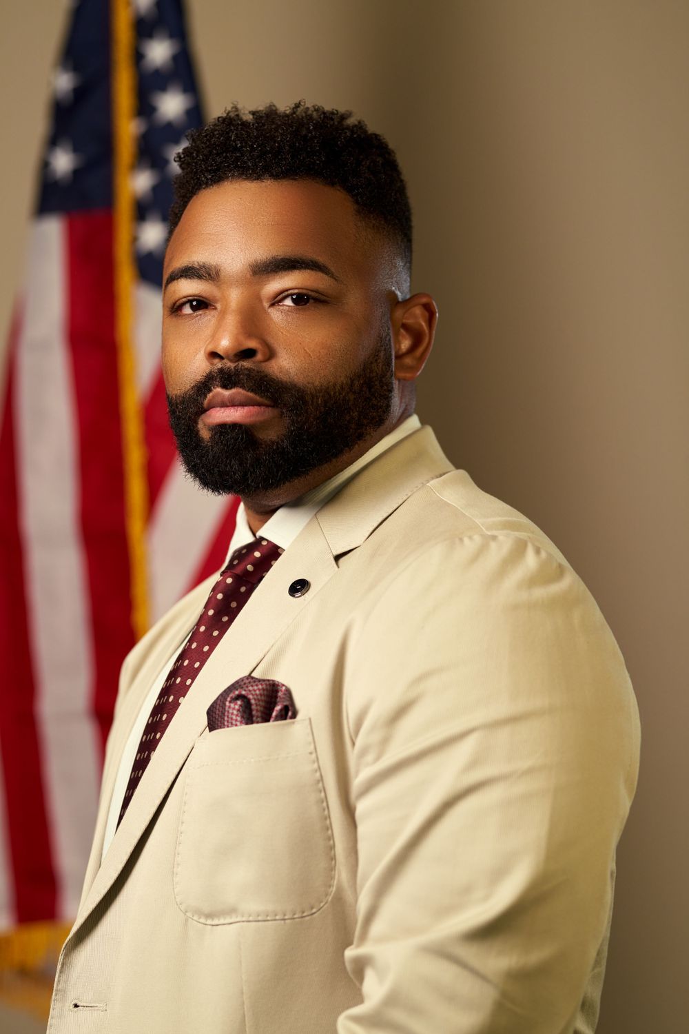 Professional portrait of official in cream suit and red tie standing before American flag.