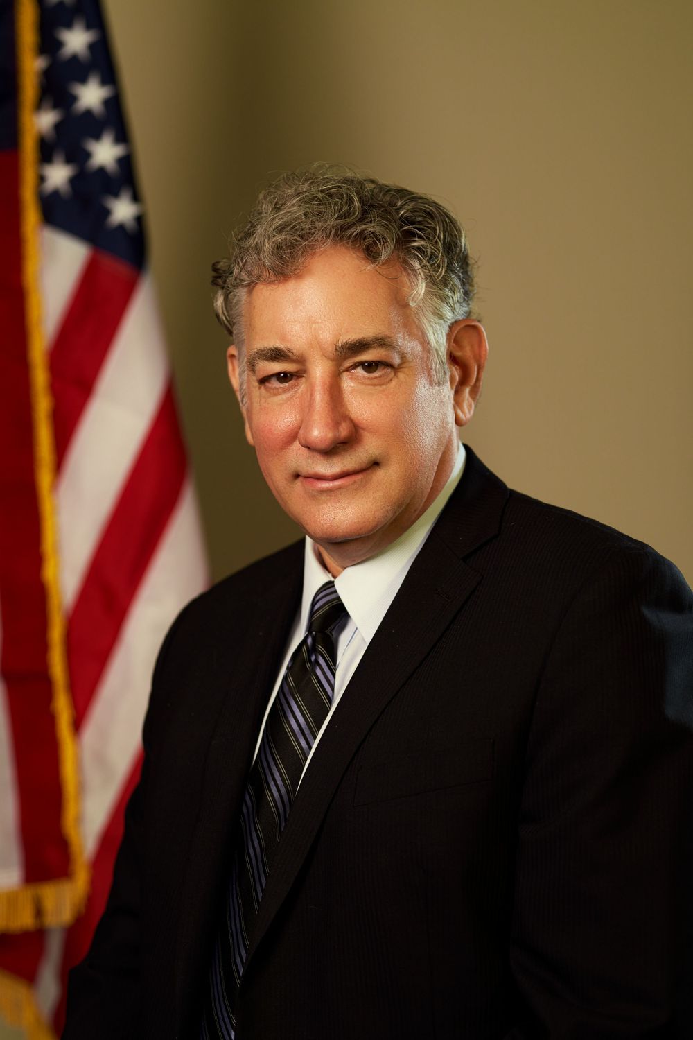 Professional headshot of senior official in black suit with striped tie against American flag backdrop.
