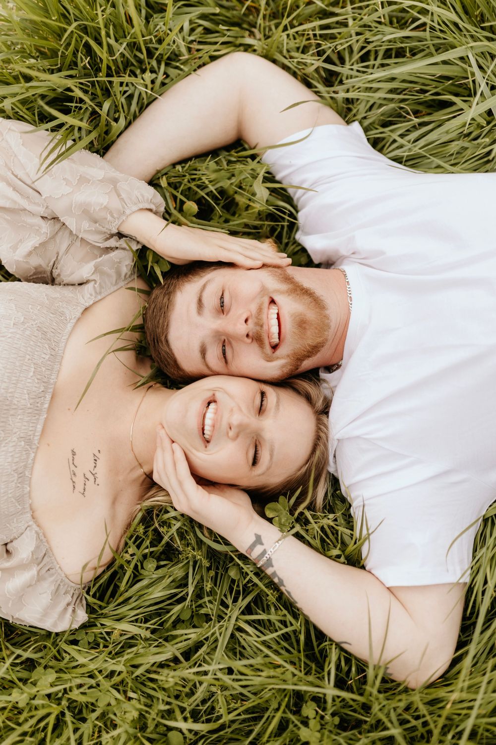 A couple lying in tall grass sharing laughter during an outdoor photoshoot.