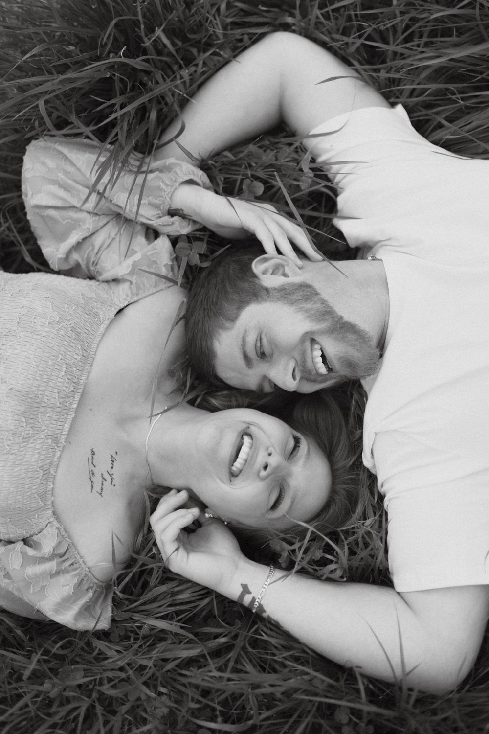 A black and white photo of a couple lying in grass sharing joyful laughter.