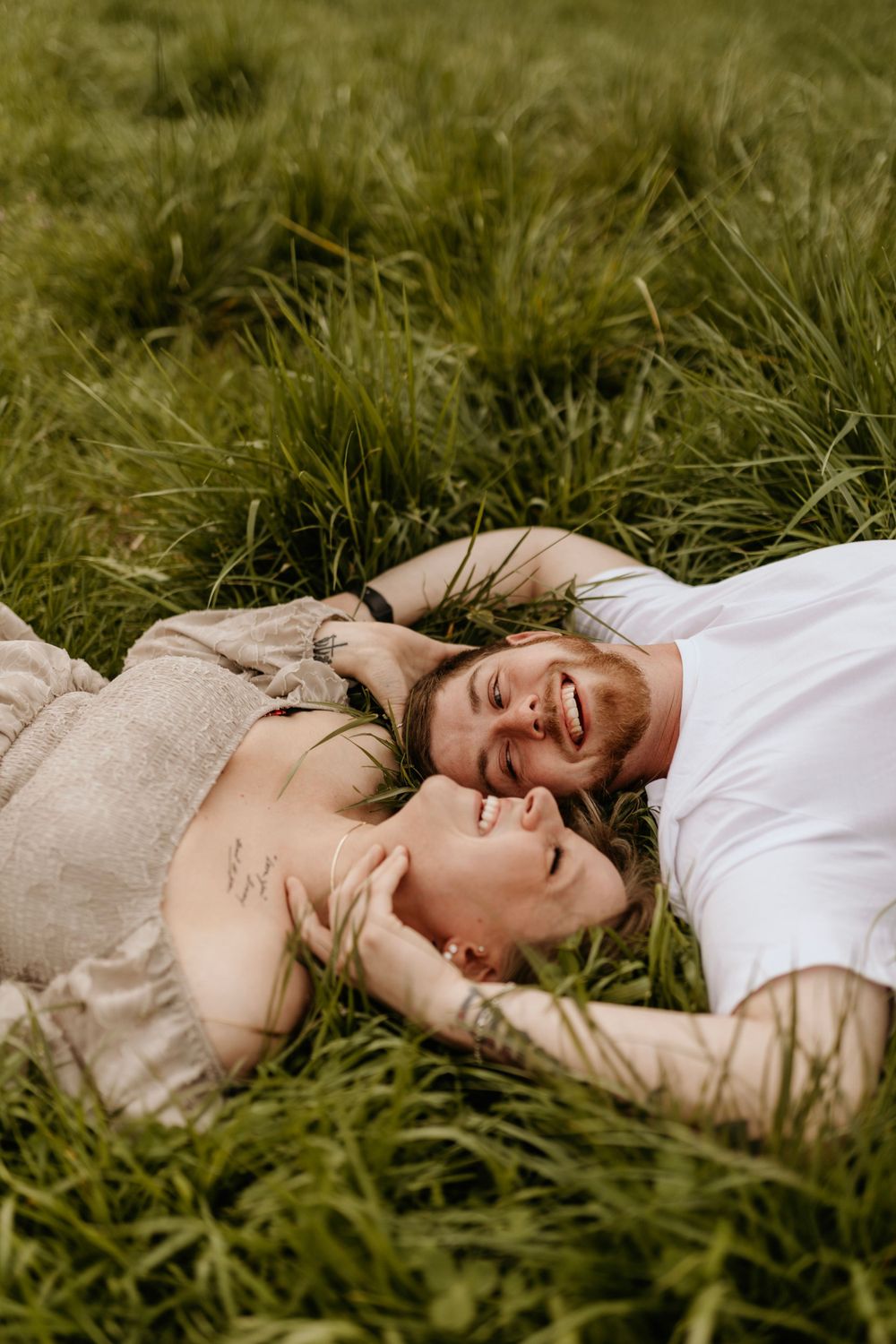 A couple laughs while lying in tall grass during a romantic outdoor photoshoot.