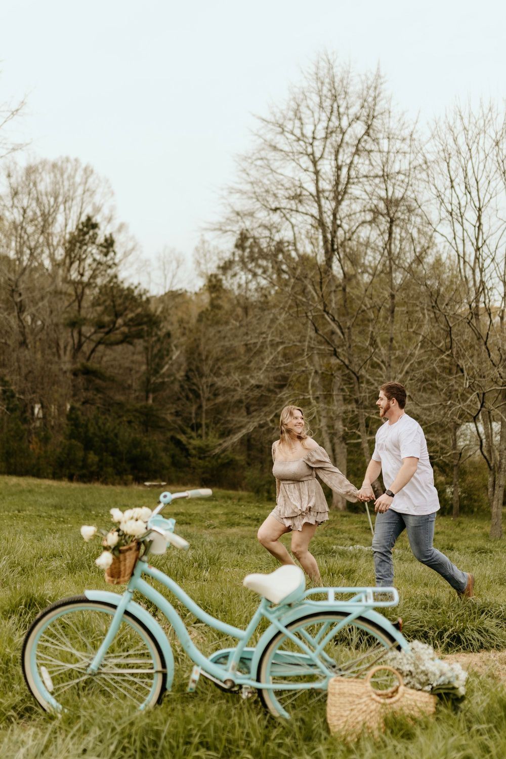 A couple runs playfully in a grassy field with a mint green vintage bicycle adorned with flowers and a basket.