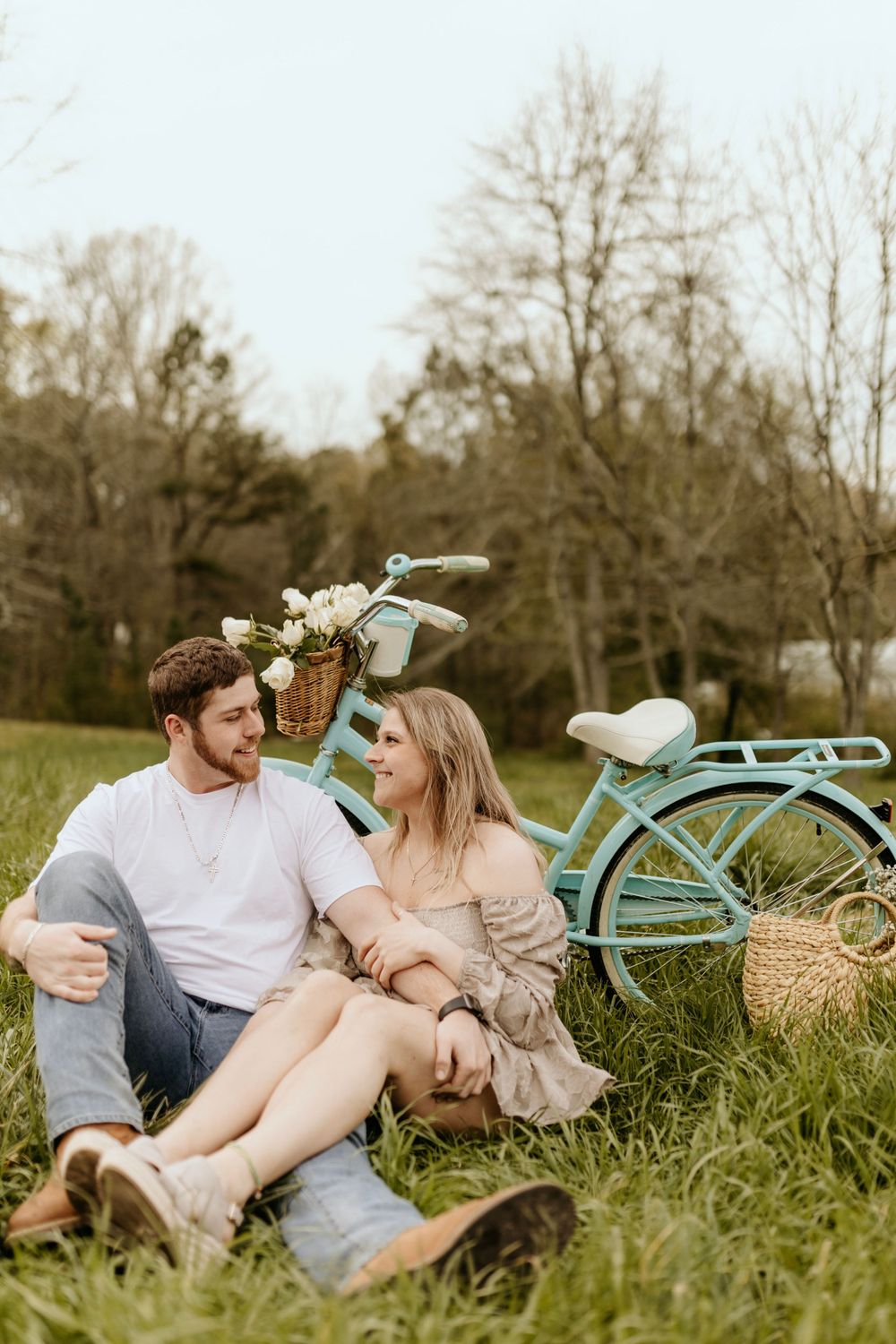 A couple relaxes on grass near a vintage mint bicycle with flowers and wicker baskets.