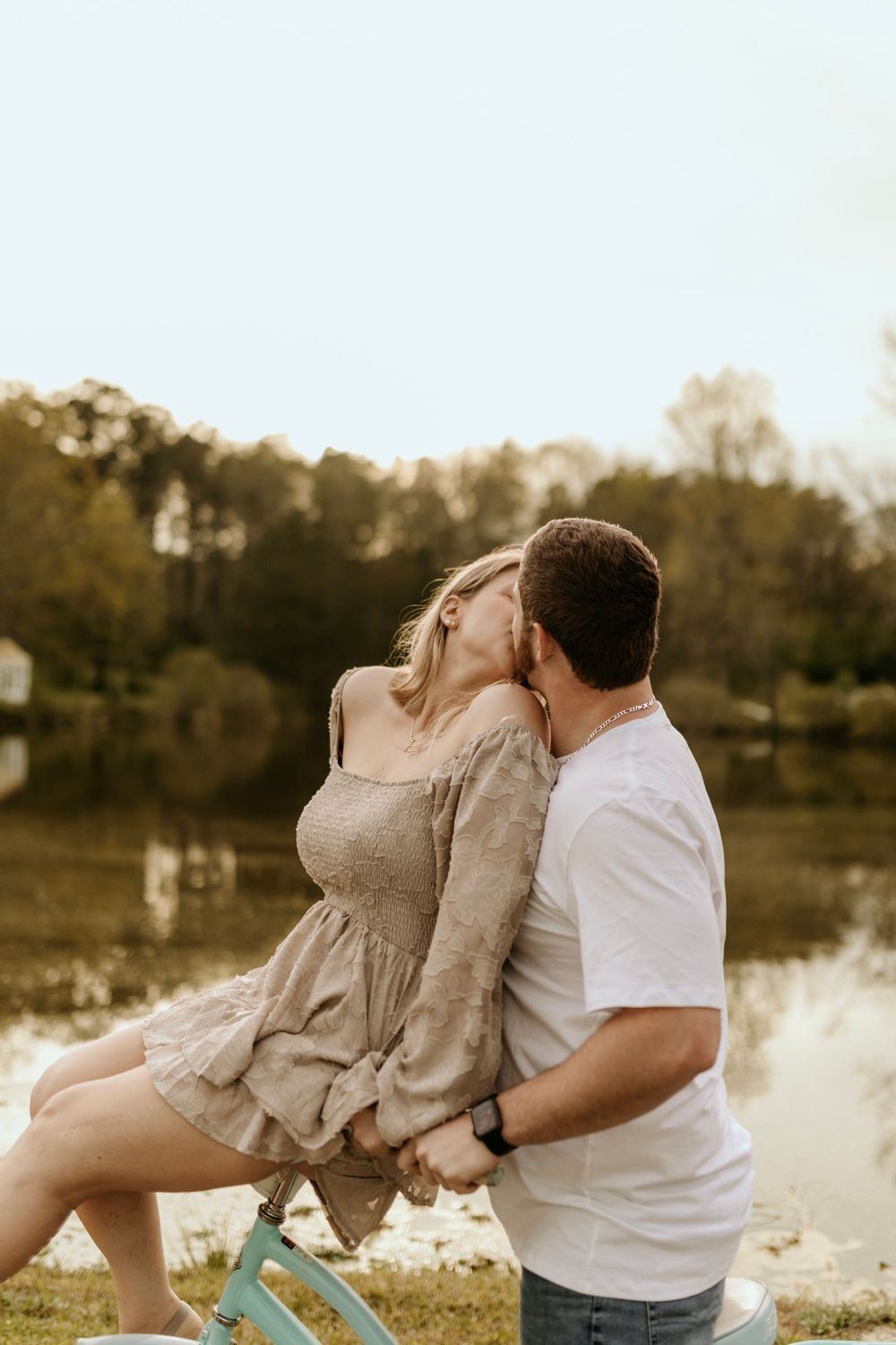 A romantic couple shares a playful moment by a lake with a mint green bicycle nearby.