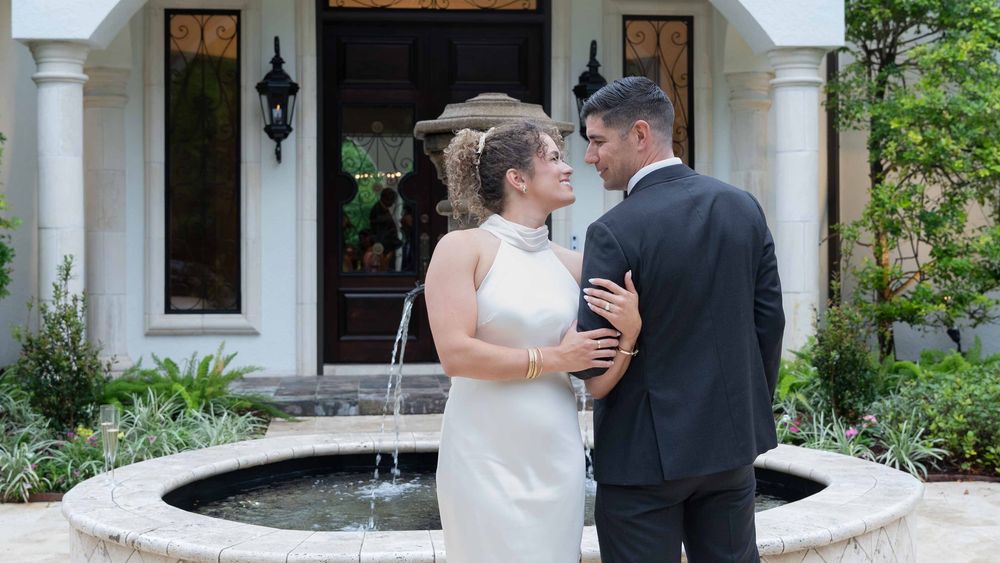 Bride and groom share romantic moment by outdoor fountain at elegant white mansion wedding venue.