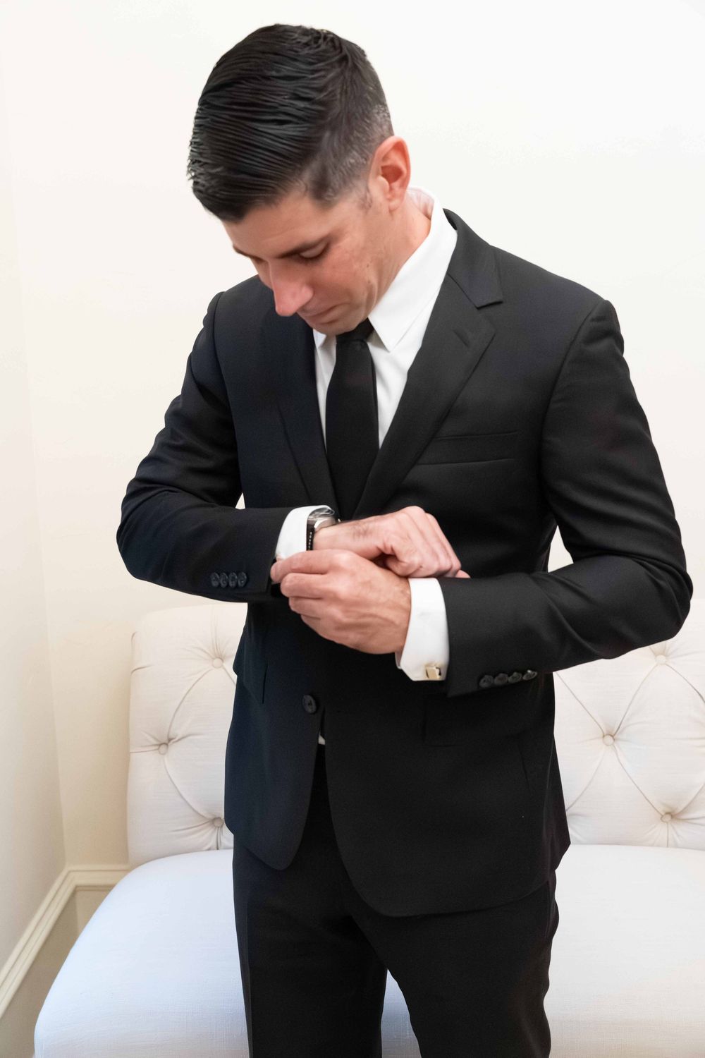 Man in black suit adjusting cufflinks, wearing white dress shirt and black tie, standing near tufted chair.