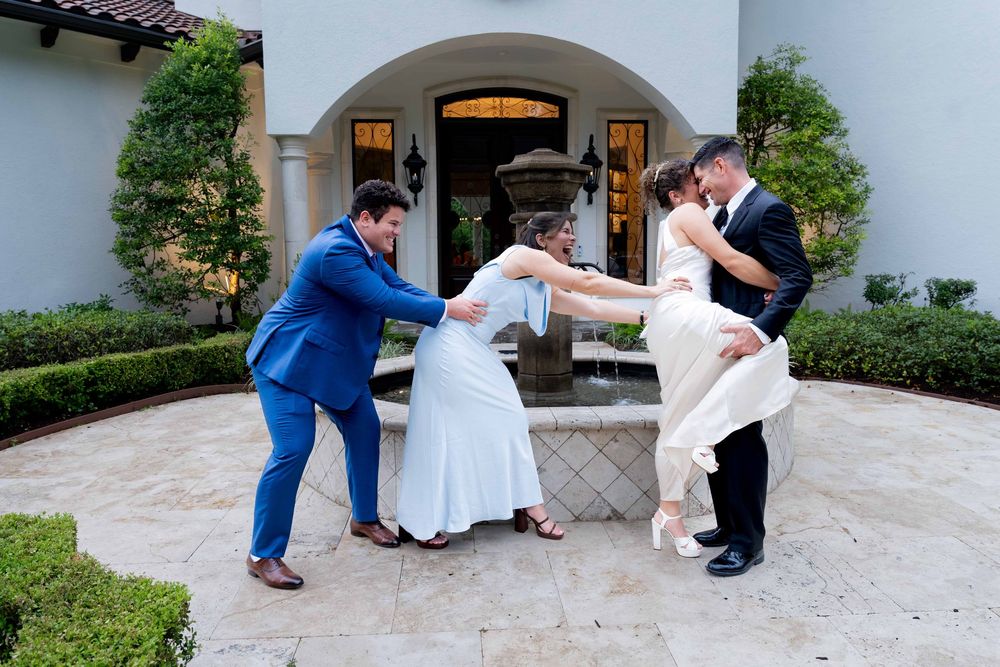 Two couples share a joyful wedding moment outside an elegant venue, with groomsmen playfully lifting brides in white gowns.
