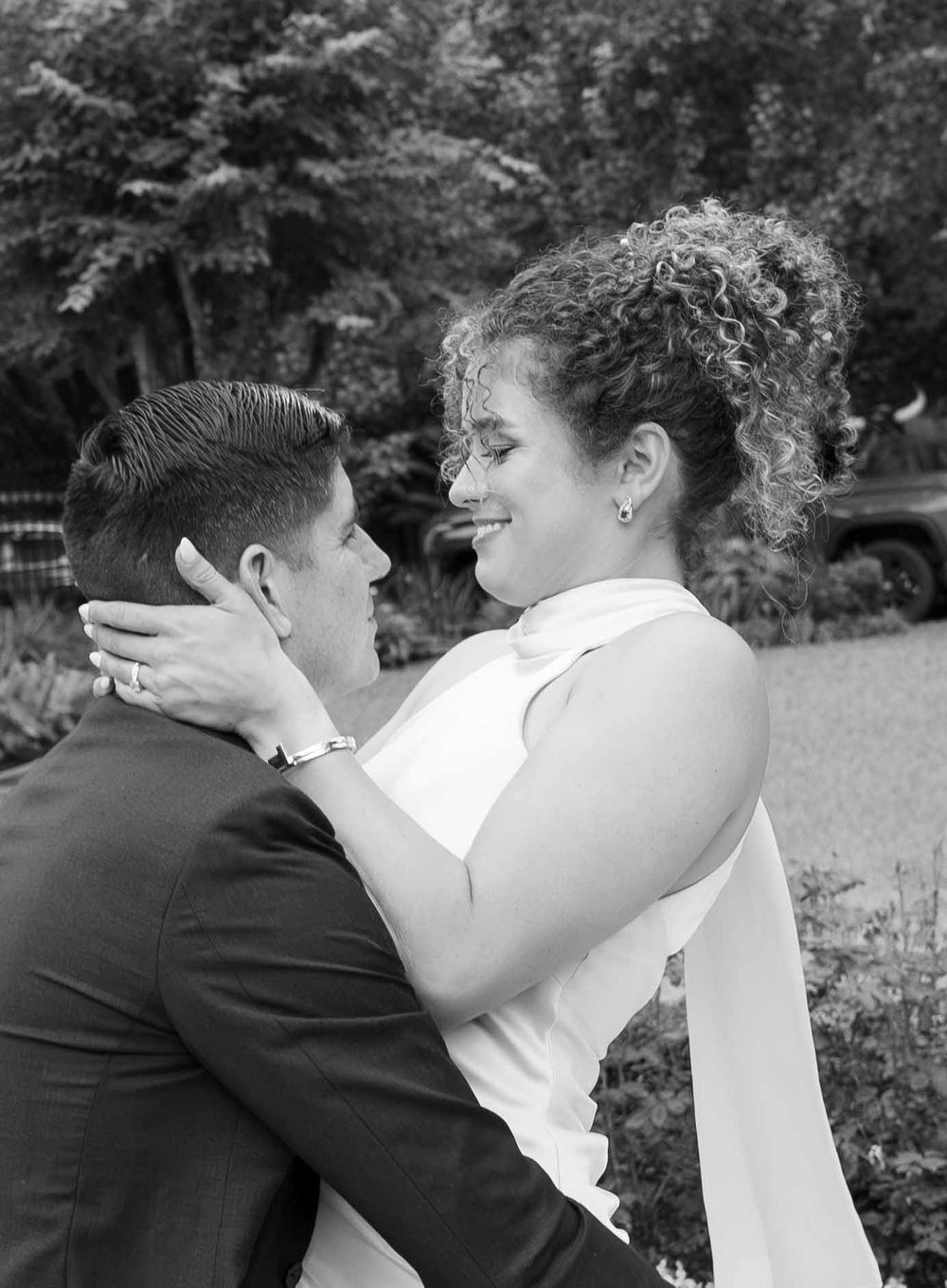 Black and white photo of a bride and groom embracing outdoors, the smiling bride in a white gown holding the groom close.
