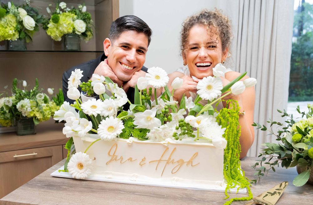 Smiling couple posing behind a white wedding cake decorated with white daisies and greenery, inscribed with Iris and High.