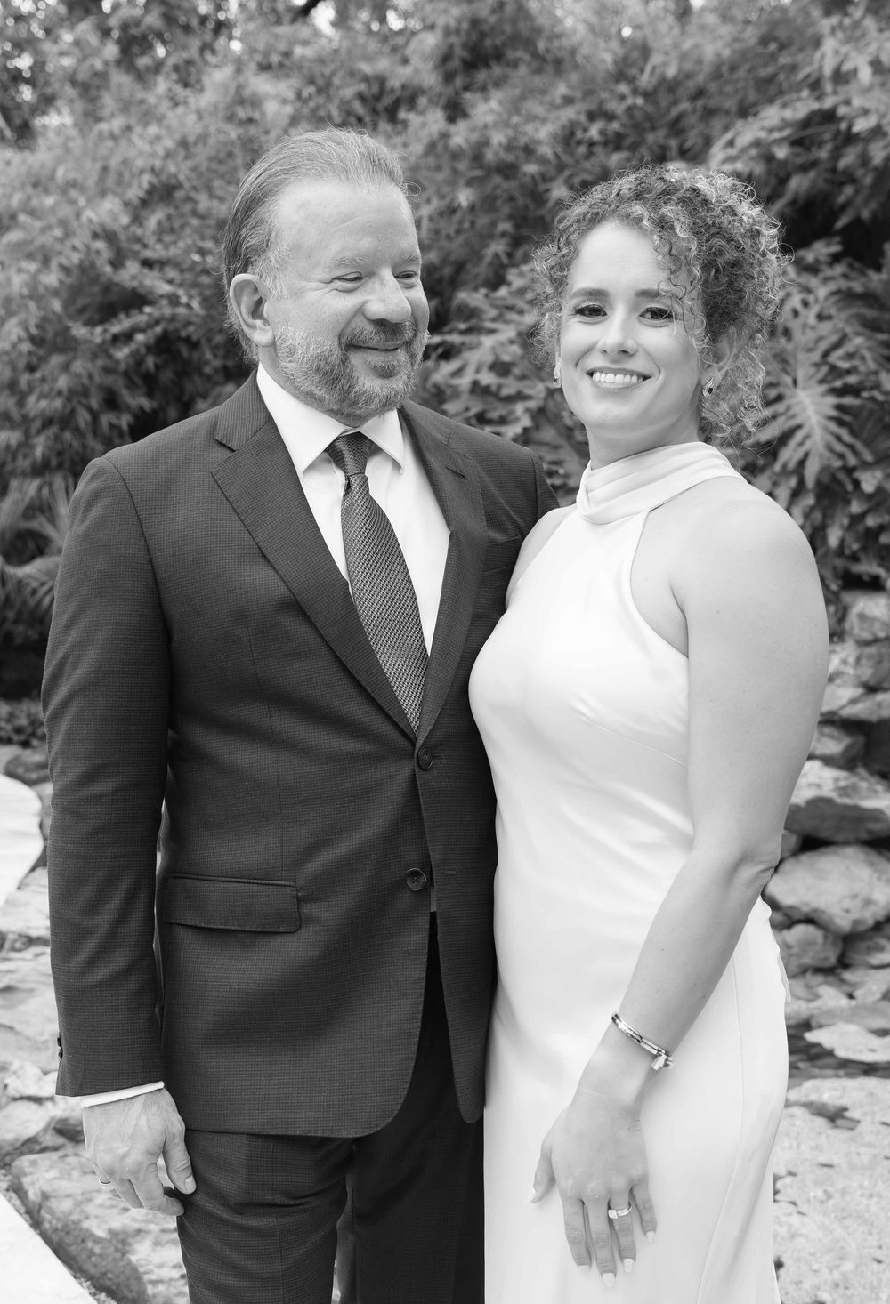 Black and white portrait of a smiling couple dressed formally, posing outdoors with lush greenery in the background.
