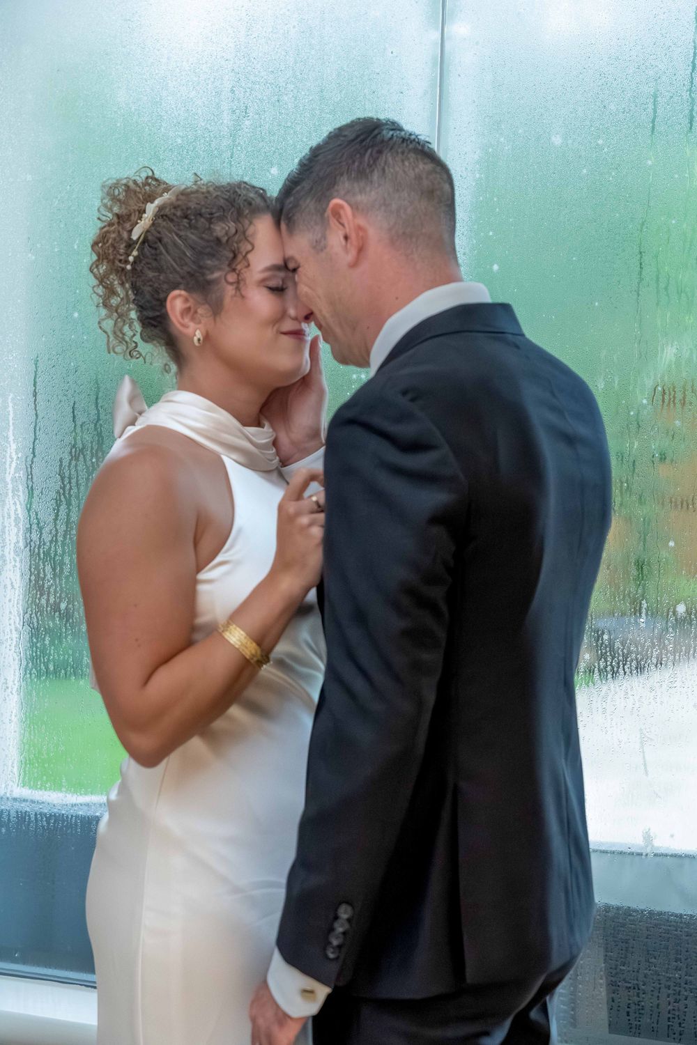 A bride and groom share an intimate moment, touching foreheads tenderly in front of a rain-covered window.