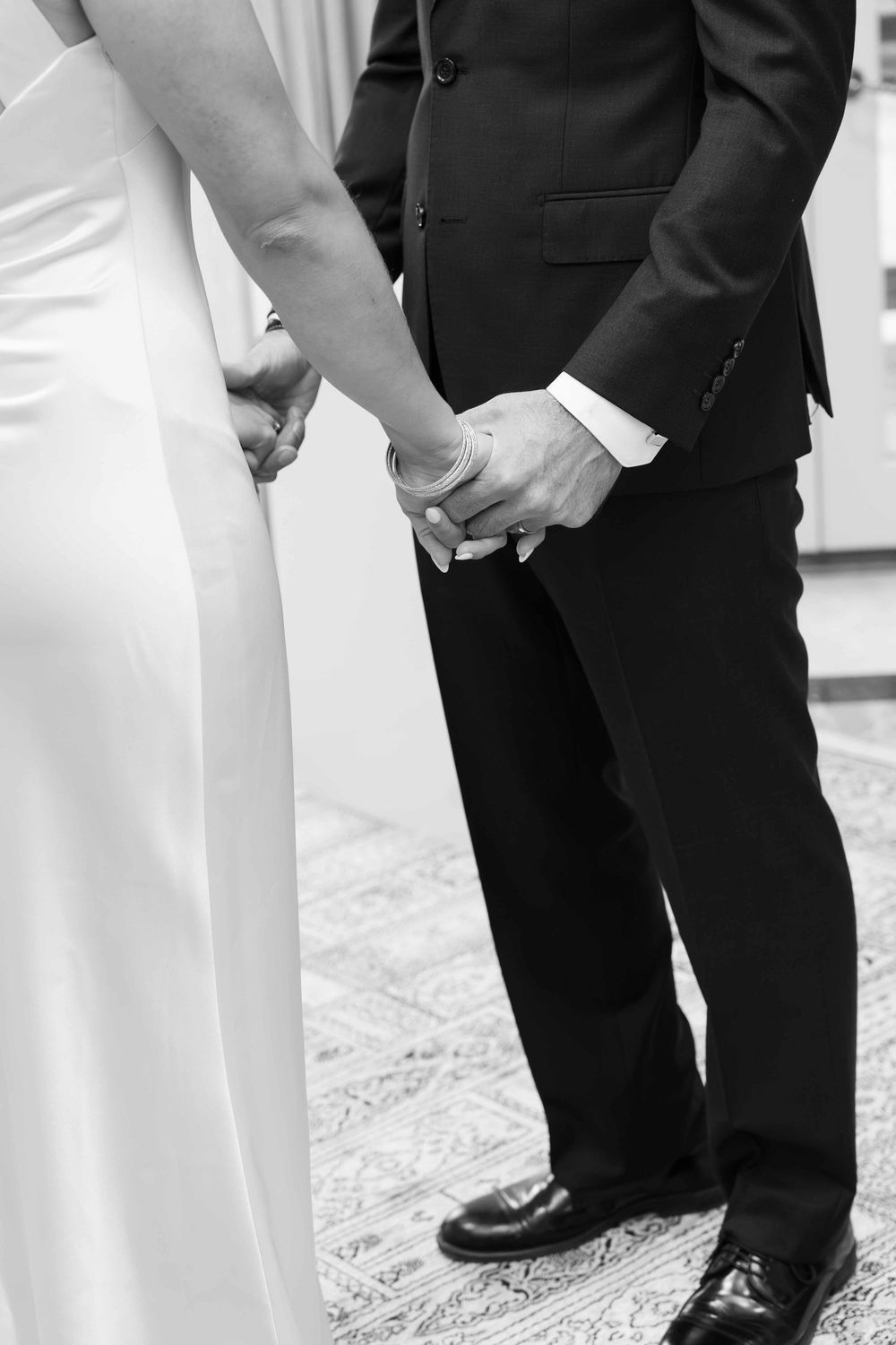 Black and white photo of bride and groom holding hands at wedding ceremony, wearing formal attire.