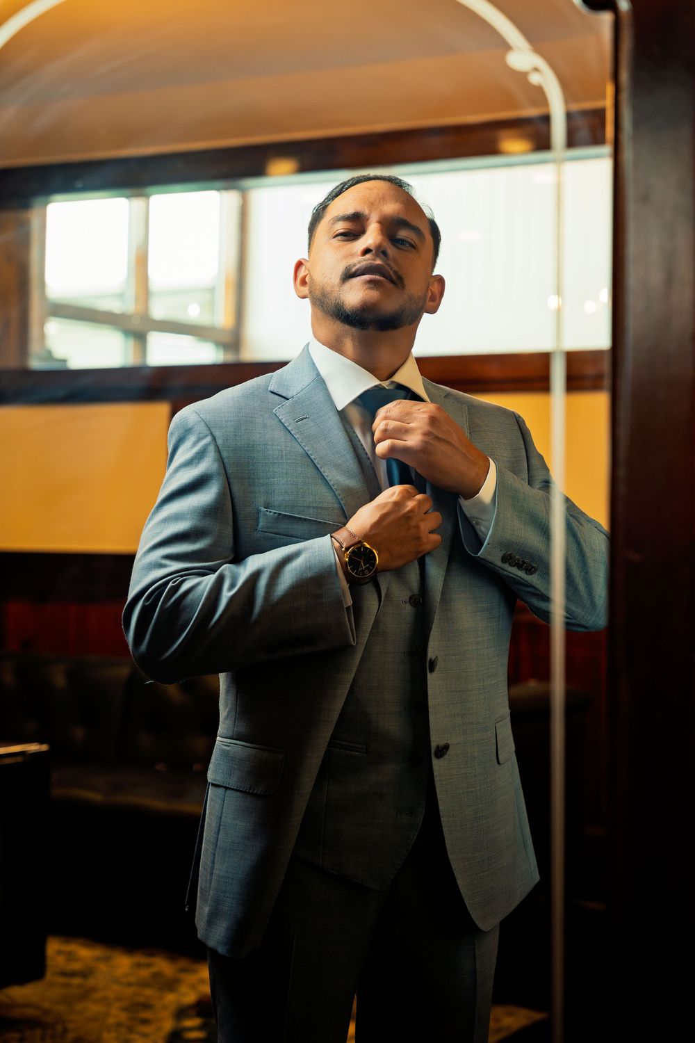 Groom in grey suit adjusting his tie while getting ready in warm indoor lighting.