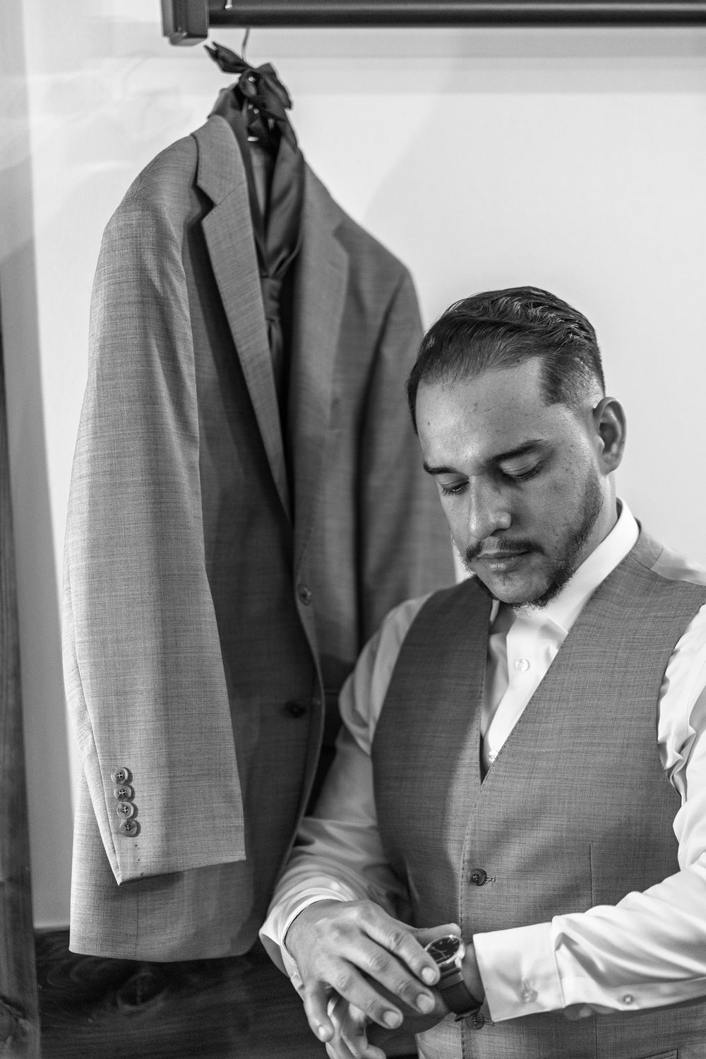 A person in a vest and white shirt checks their watch while standing next to a hanging suit jacket.