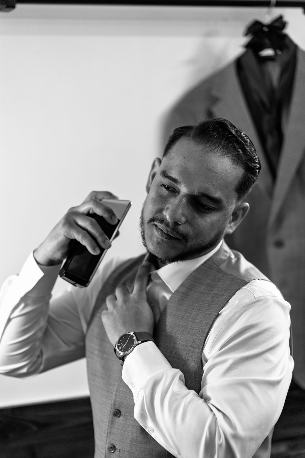 A man in a white dress shirt and grey vest adjusts his tie while getting ready.
