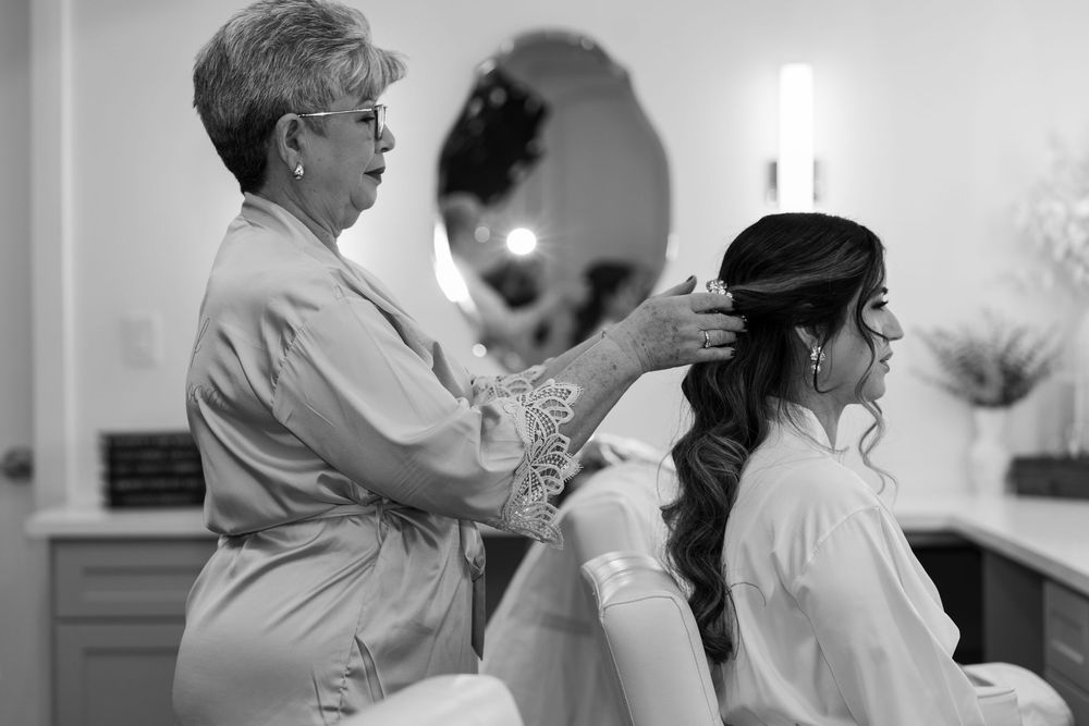 A stylist arranges a bride's hair in a salon with a round mirror visible in the background.