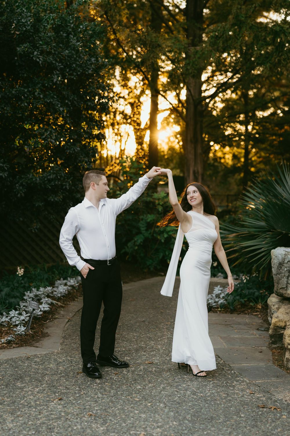 A couple dances together on a garden path at sunset with golden light filtering through trees.