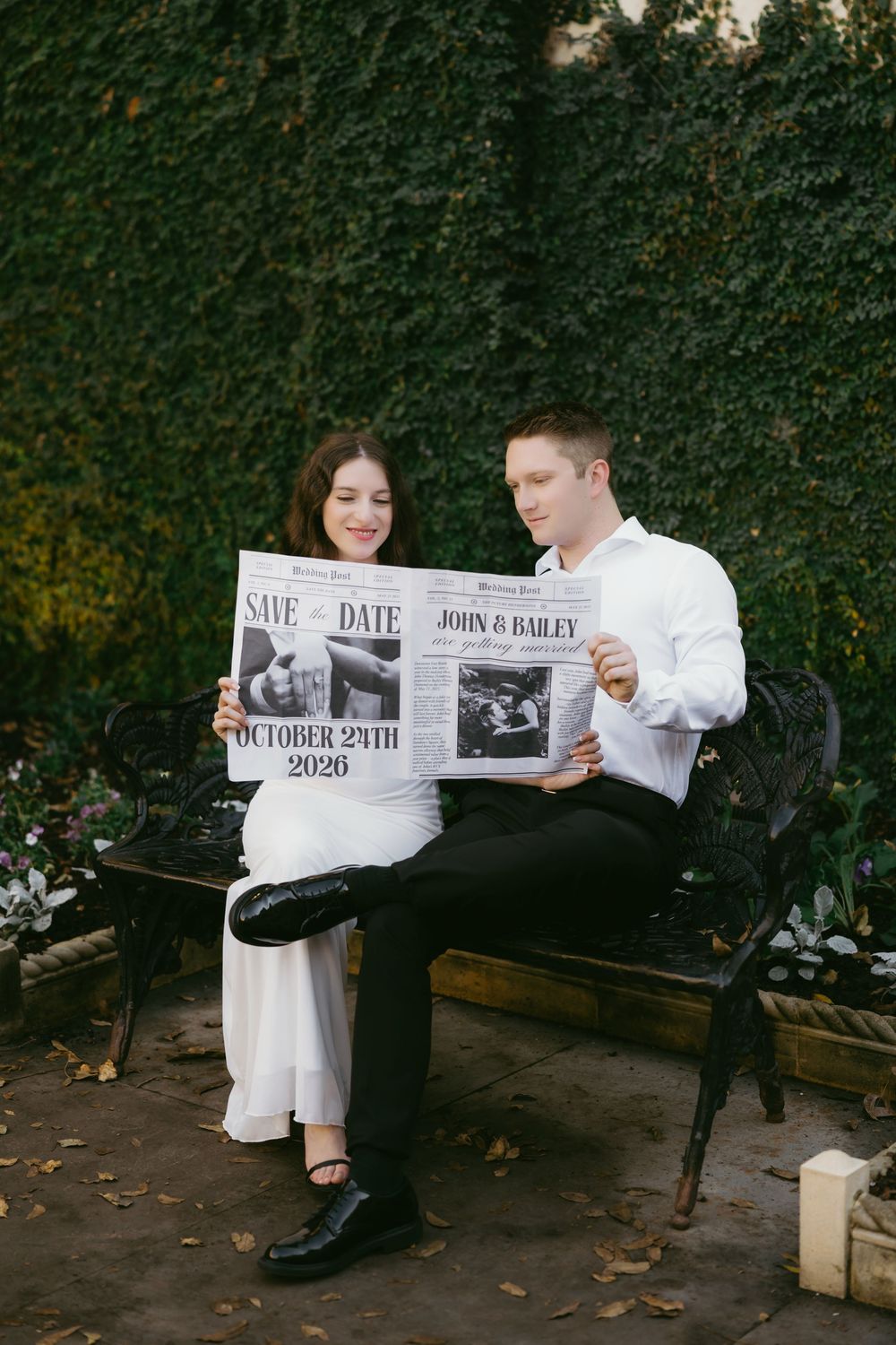 A couple sits on a bench holding a newspaper-style save the date announcement against an ivy-covered wall background.