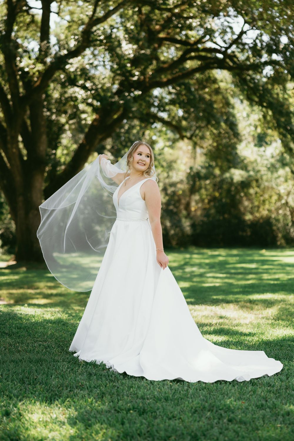 A bride in a flowing white wedding gown sits gracefully on lush green grass under the dappled shade of trees.