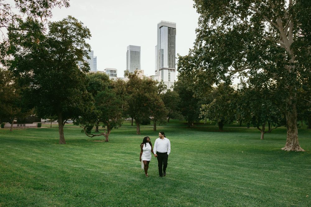 Two people walk through a lush green park with tall trees and city buildings visible in the background on a sunny day.