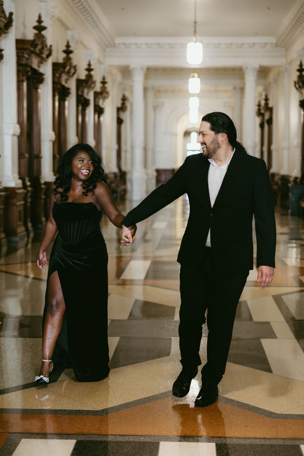 Elegant couple in formal attire holding hands while walking down ornate hallway with vintage architectural details.