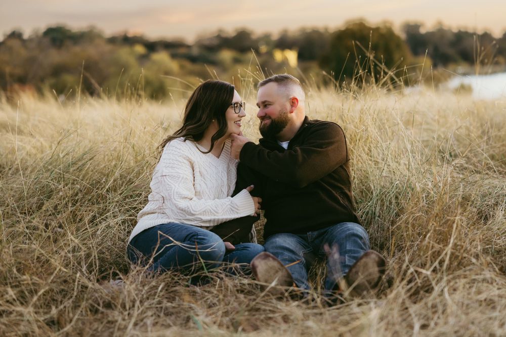 A couple sits together in a golden field of tall grass during sunset, sharing an intimate moment.