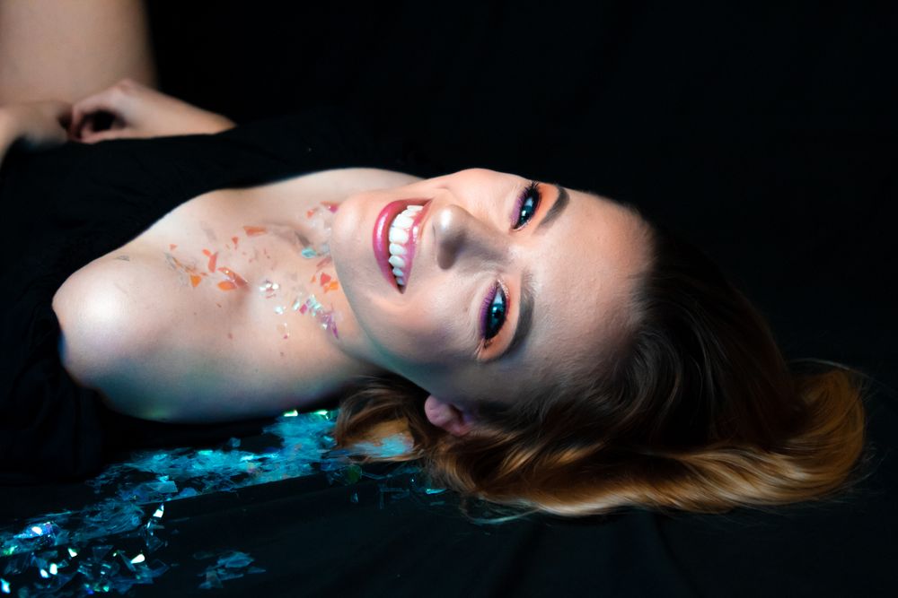 A person wearing pink and orange makeup laying on black fabric with glitter looking at the camera.