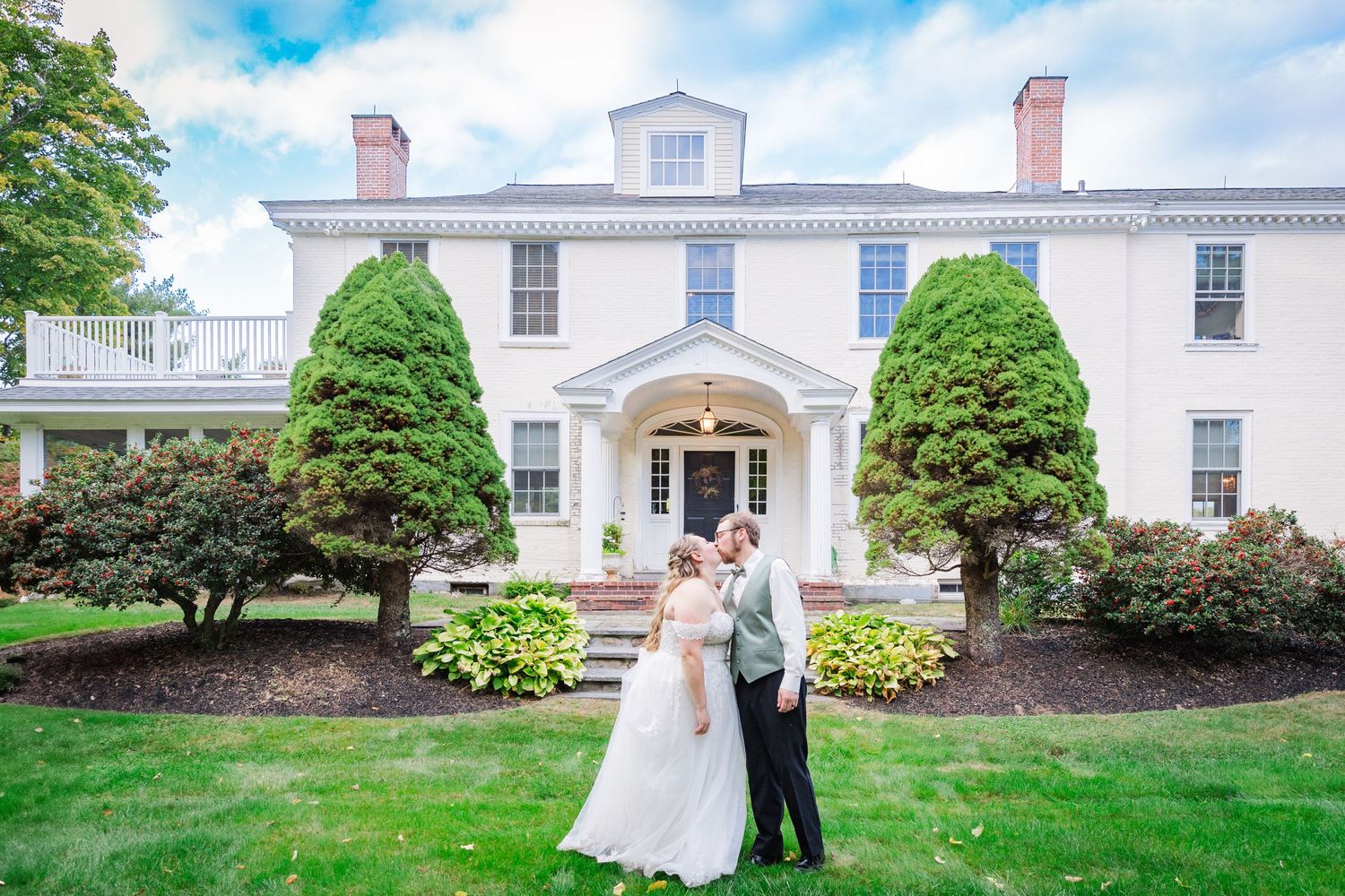 An elegant wedding couple stands in front of a grand white colonial mansion with manicured landscaping.